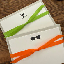 2 sets of cards, one with a skier silhouette and one with a pair of black sunglasses on  each pack tied with either 
an orange and a green ribbon