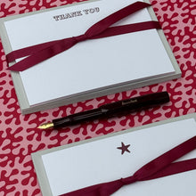 A gift set featuring two white envelopes tied with burgundy ribbons, one printed with 'THANK YOU' and the other with a small star, accompanied by a black fountain pen with a gold nib, all arranged on a red and white patterned background.
