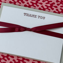 A white square card with the words 'THANK YOU' printed in a dark serif font, wrapped with a dark red ribbon bow, placed on a pink and red abstract patterned background.