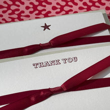 A white card with 'THANK YOU' and a small star, tied with a dark red ribbon, placed on a red-and-white patterned surface with a dark red pen at the bottom.