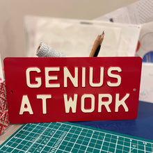 Red metal sign with light cream text saying Genius At Work
