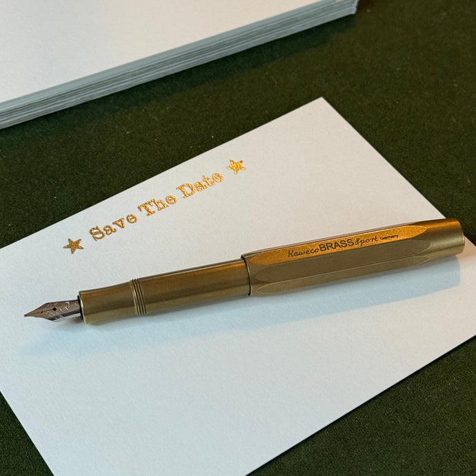 A solid brass fountain pen on a white postcard style notecard with the text Save The Date